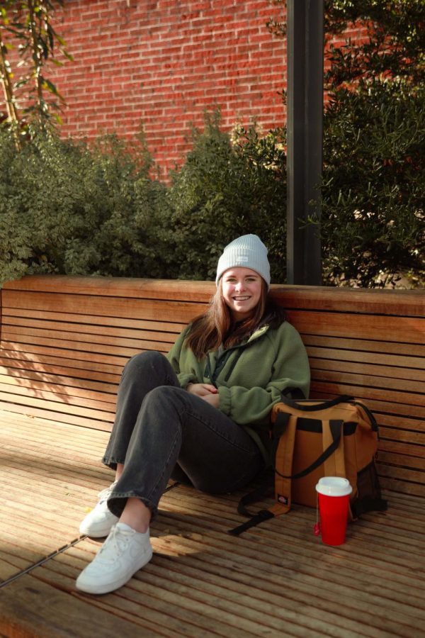 McKenzie Scott sits on a bench next to a backpack and a hot cup of tea while wearing a beanie.