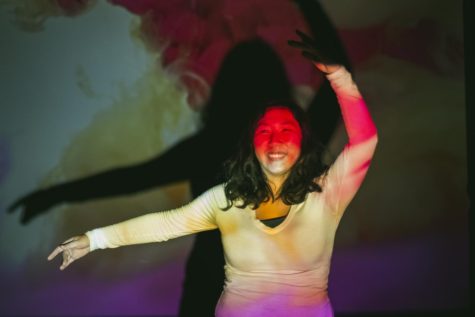 Julianna Souther is laughing and dancing with hands above head. A projector is projecting a pink and yellow light across Souther and the background.