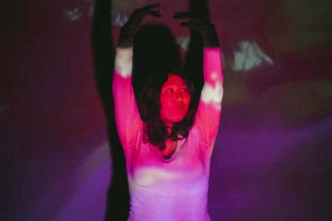 Julianna Souther is dancing with hands above head. A projector is projecting a pink light across Souther and the background.