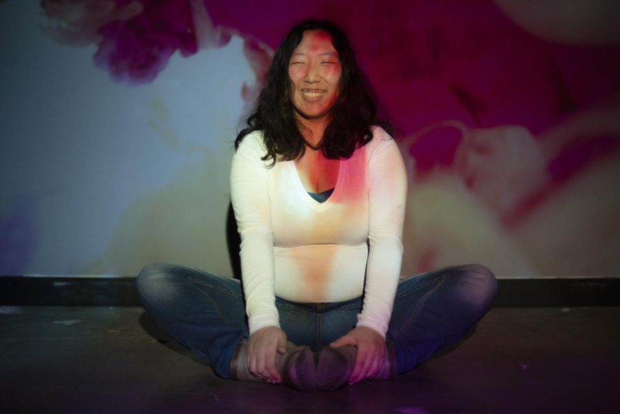 Julianna Souther is sitting on the floor laughing. A projector is projecting a pink and yellow light across Souther and the background.