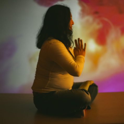 Julianna Souther meditates, but the photo is of Souther's side profile, so only their right side is showing. A projector is projecting a pink light across Souther and the background.