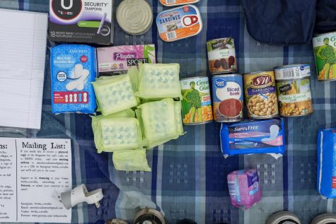 A table covered with pads, tampons, notebook paper, and canned foods such as sardines, sliced beets, sweet corn and chickpeas.
