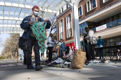 Donovon Horst wears a t-shirt, denim jacket, denim jeans, and a mask as he walks away from the RRFM with a zine and clothing items in-hand. On article of clothing features a strawberry print.