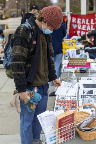 Ian Hermanson wears a knit beanie, mask, a zip-up jacket and a printed sweater over top, and classic denim jeans, as he flips through a handmade artist zine with one hand, with several clothing items in the other hand.