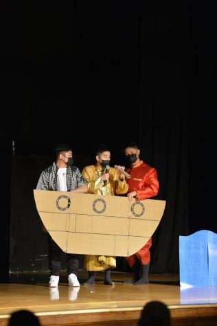 Christian Her, left, Aaren Kong and Austin Peng, right, stand together on stage. Ittihrit speaks into a microphone while the other two hold up a boat prop made of cardboard.