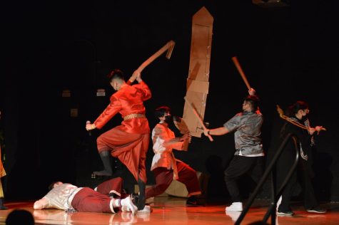 A group of students act out a fight scene with sword props made of cardboard on stage.
