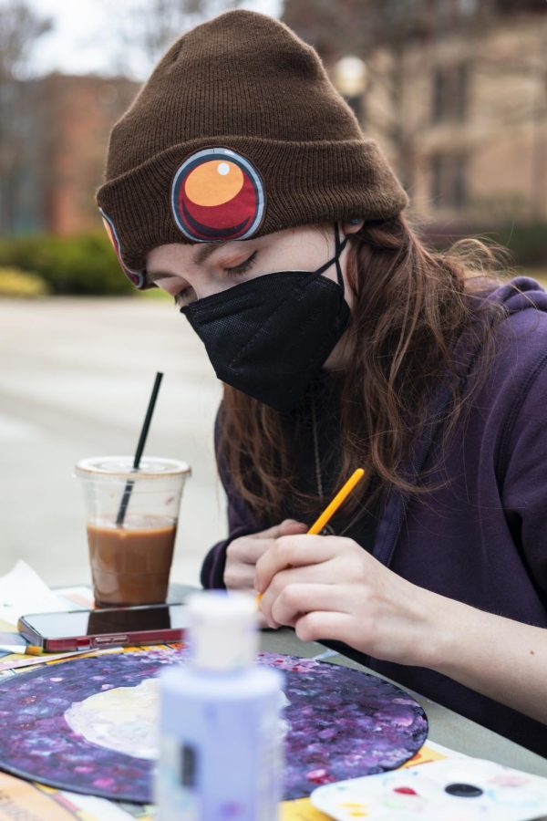 Sage Derting wears at sweatshirt and beanie , an iced coffee at her side, as she paints a record.