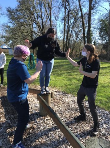 One person walks along a balance beam while two people, one on each side of the balancer, help guide them across the beam.