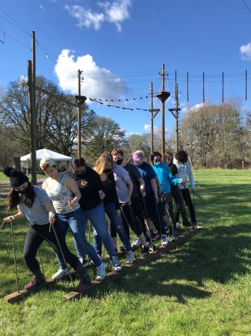 A group of people stand on a contraption that resembles an 11-person ski. They must coordinate their steps so they can move forward collectively.