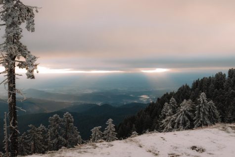 The photo looks over a snowy cliff to reveal a view of trees and hills below. The sky is pink and blue and the sun is rising.